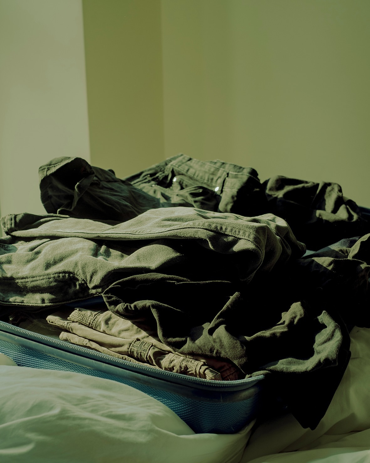 Open blue suitcase filled with folded clothing on a white bed in a brightly lit room with cream colored walls.