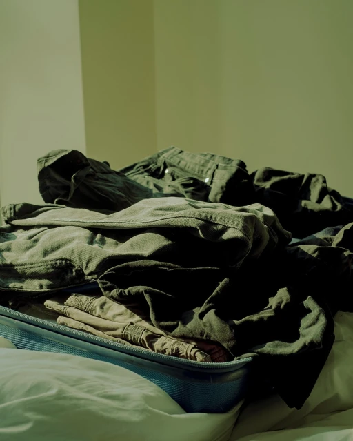 Open blue suitcase filled with folded clothing on a white bed in a brightly lit room with cream colored walls.