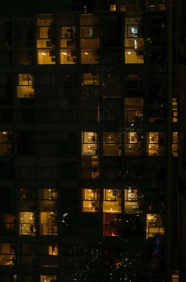 Exterior vertical nighttime view of a tall apartment building with windows lit from within, creating a warm glow against the dark sky.