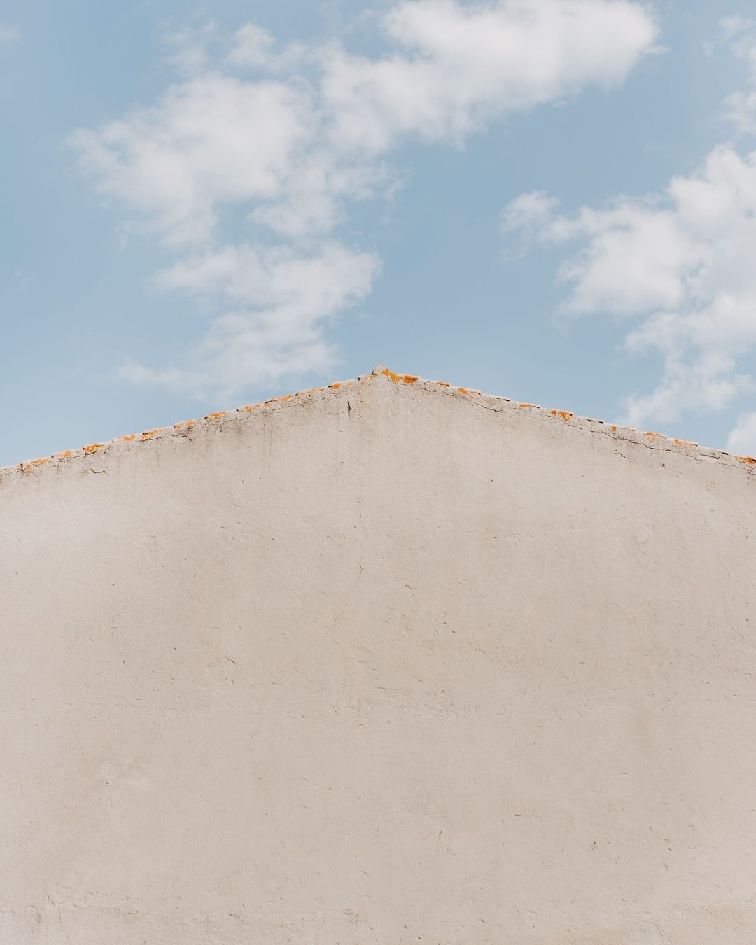 Abstract shot of a building facade with white stucco under a pale blue sky with scattered clouds in the background, daytime.