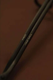 Close-up of black wick trimmer with white text reading BYREDO and L'OBJET SIMPLE against a dark brown background