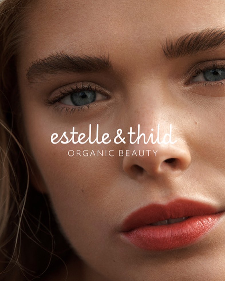 Close-up of a woman's face with blue eyes and red lips. Text overlay reads: estelle & thild ORGANIC BEAUTY, in a beauty product advertisement.