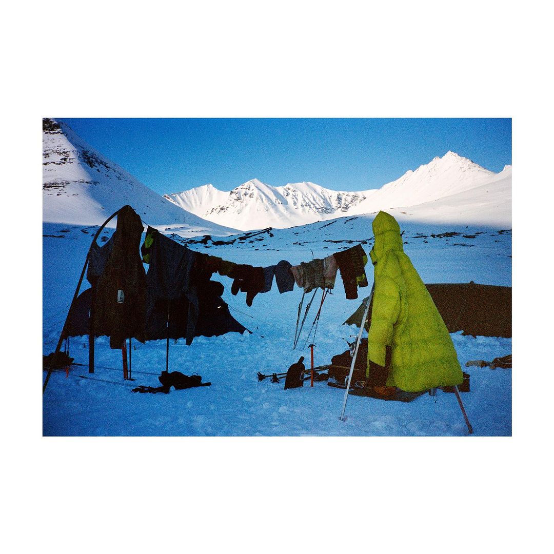 Camping scene in a snow-covered landscape under a clear blue sky, featuring a tent, hanging clothes, and towering snow-capped mountains.