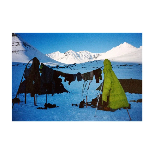 Camping scene in a snow-covered landscape under a clear blue sky, featuring a tent, hanging clothes, and towering snow-capped mountains.
