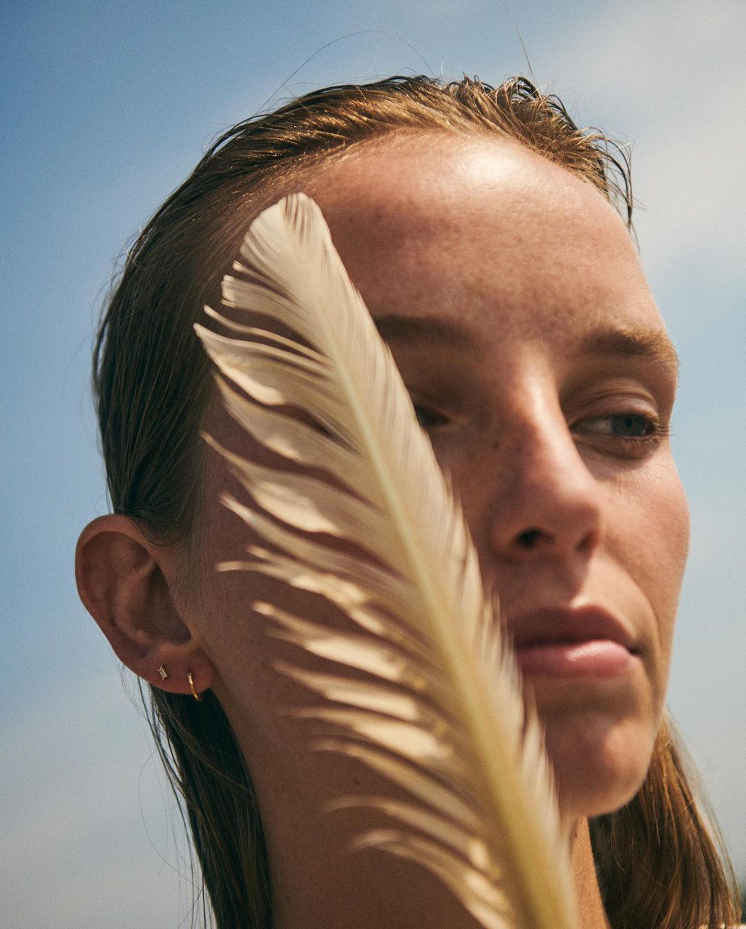 Close-up portrait of a woman with a feather in front of her face, set against a blue sky background. Gold hoop earrings are visible.