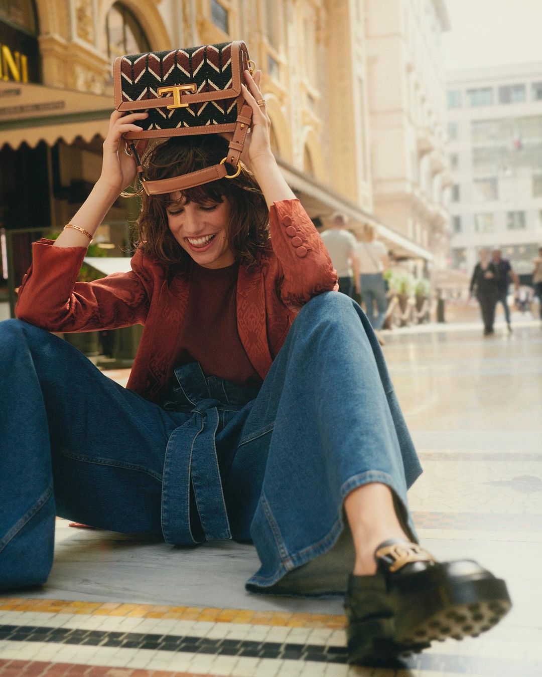 A happy woman with curly hair sits outdoors wearing wide-leg jeans, a blazer, and chunky loafers while holding a purse over her head.