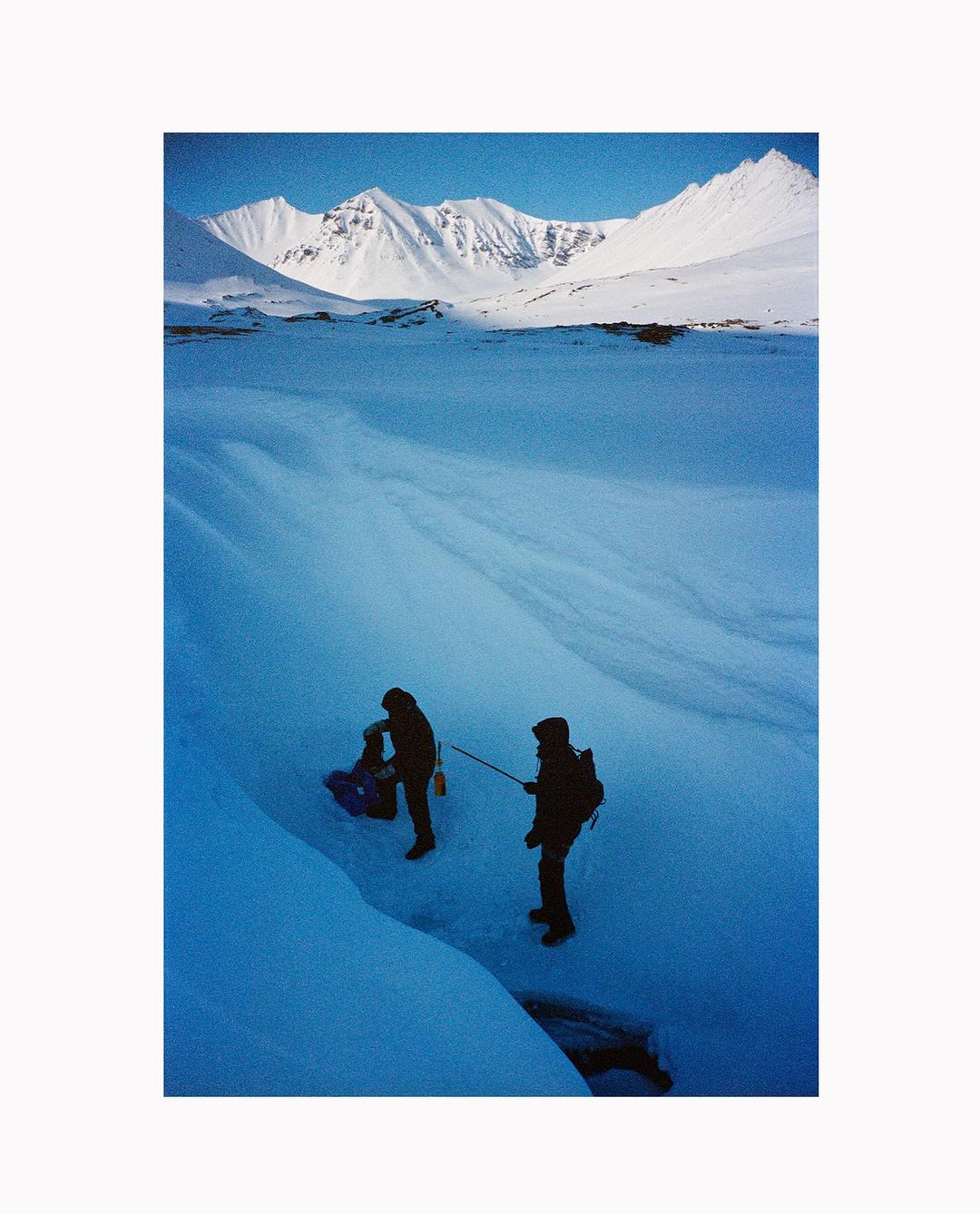 Two hikers haul a sled across a snowy, icy field towards snow-capped mountains under a clear blue sky in an outdoor landscape shot.