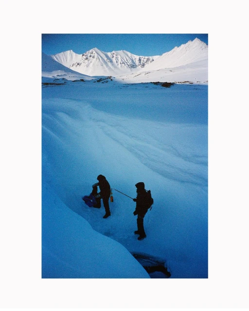 Two hikers haul a sled across a snowy, icy field towards snow-capped mountains under a clear blue sky in an outdoor landscape shot.