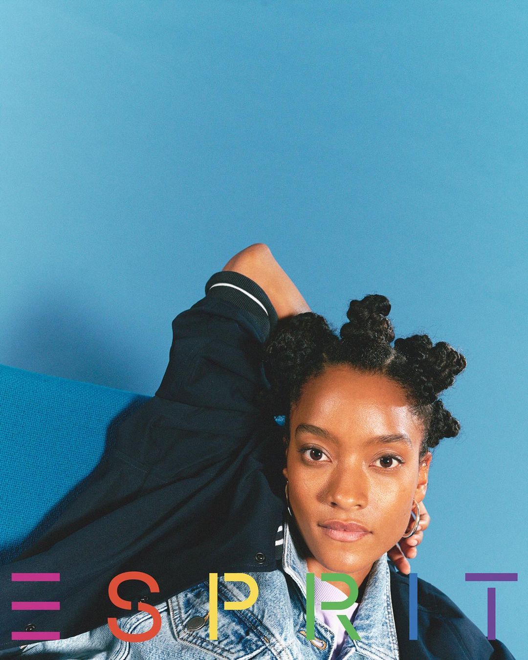 Portrait of a stylish young Black woman with a denim jacket and unique hairstyle, posing against a vibrant blue backdrop, fashion advertisement.