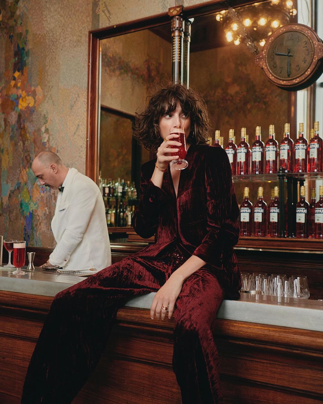 Woman in a burgundy velvet suit sits on a bar counter drinking a red cocktail; barman in background. Bottled liquor in the background.