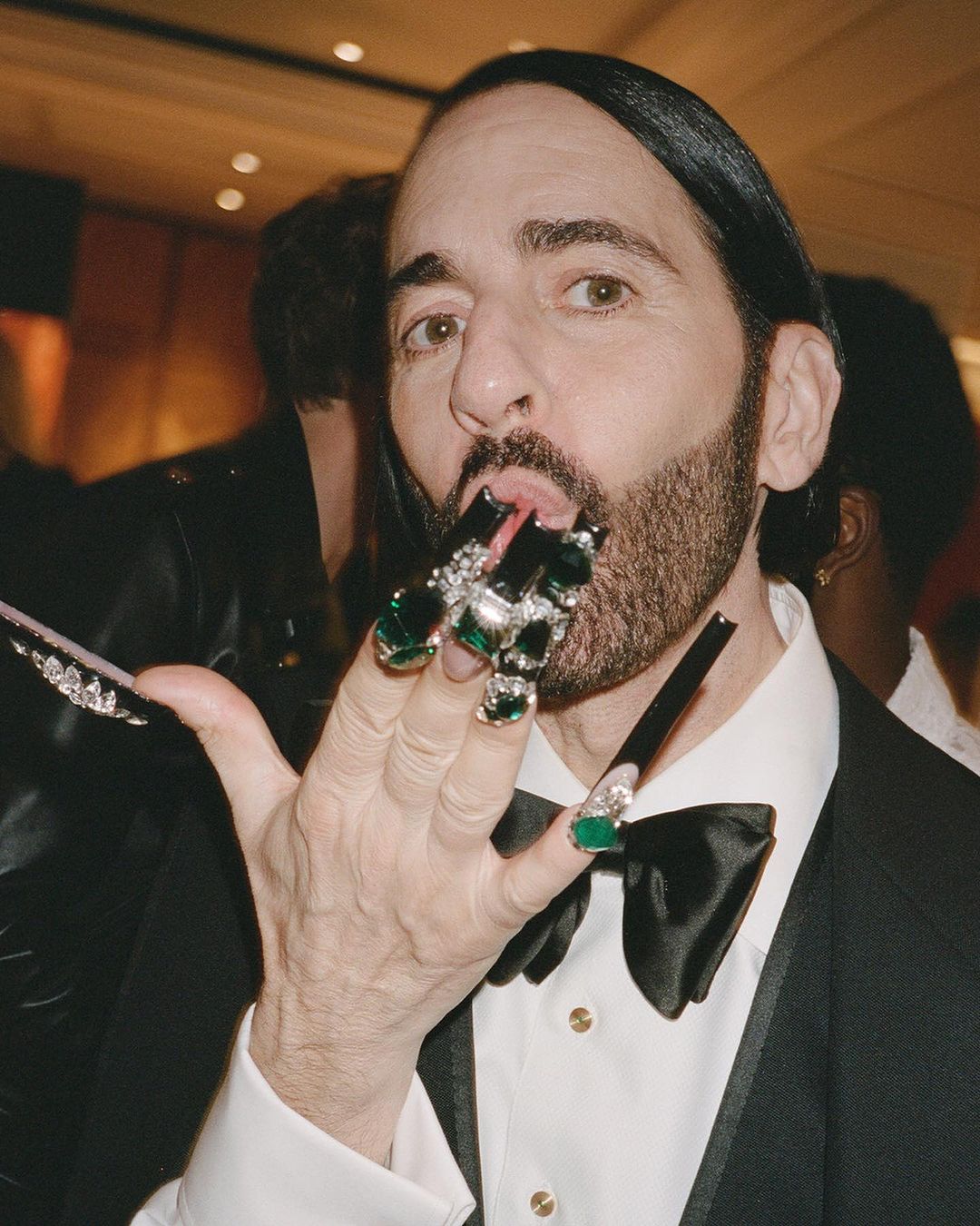 Close-up of a person with unique manicure and jewelry holding fingers near mouth, wearing tuxedo with bow tie, indoors.