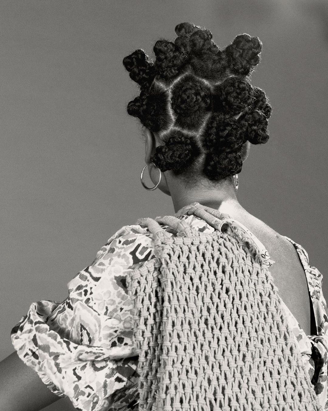 Back view of an adult female with hair styled in knot buns, wearing statement earrings, and a woven macrame bag over her shoulder.