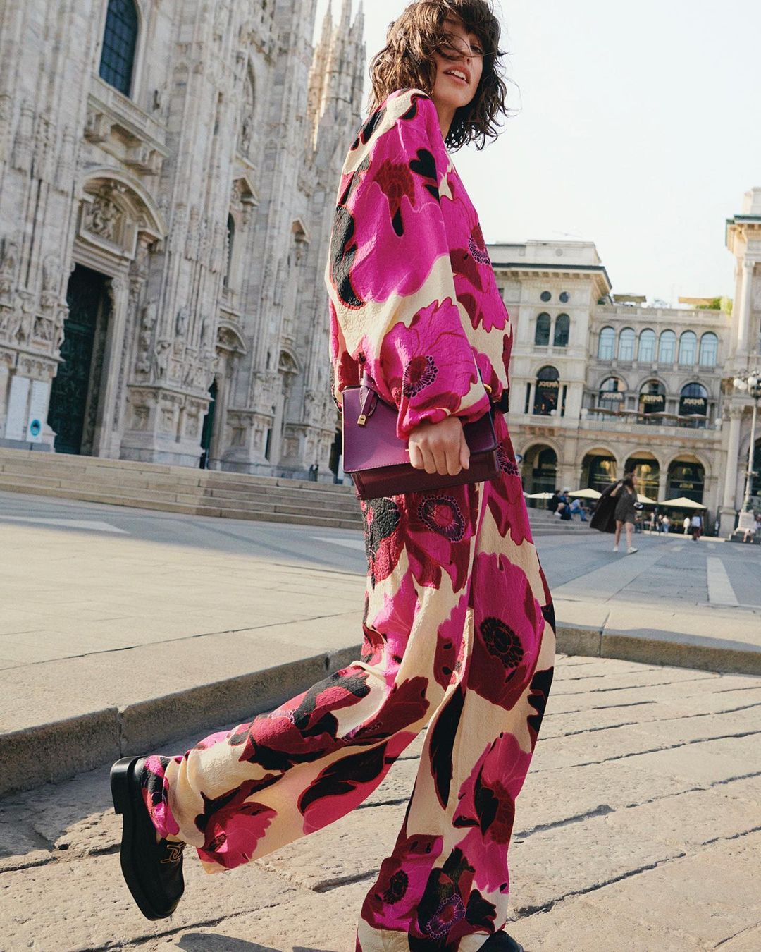 A young woman walks across a plaza wearing a matching pink floral outfit, with a cathedral in the background on a sunny day.