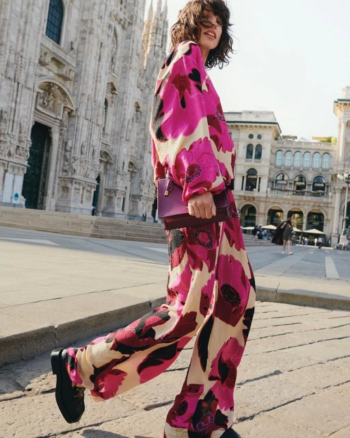 A young woman walks across a plaza wearing a matching pink floral outfit, with a cathedral in the background on a sunny day.