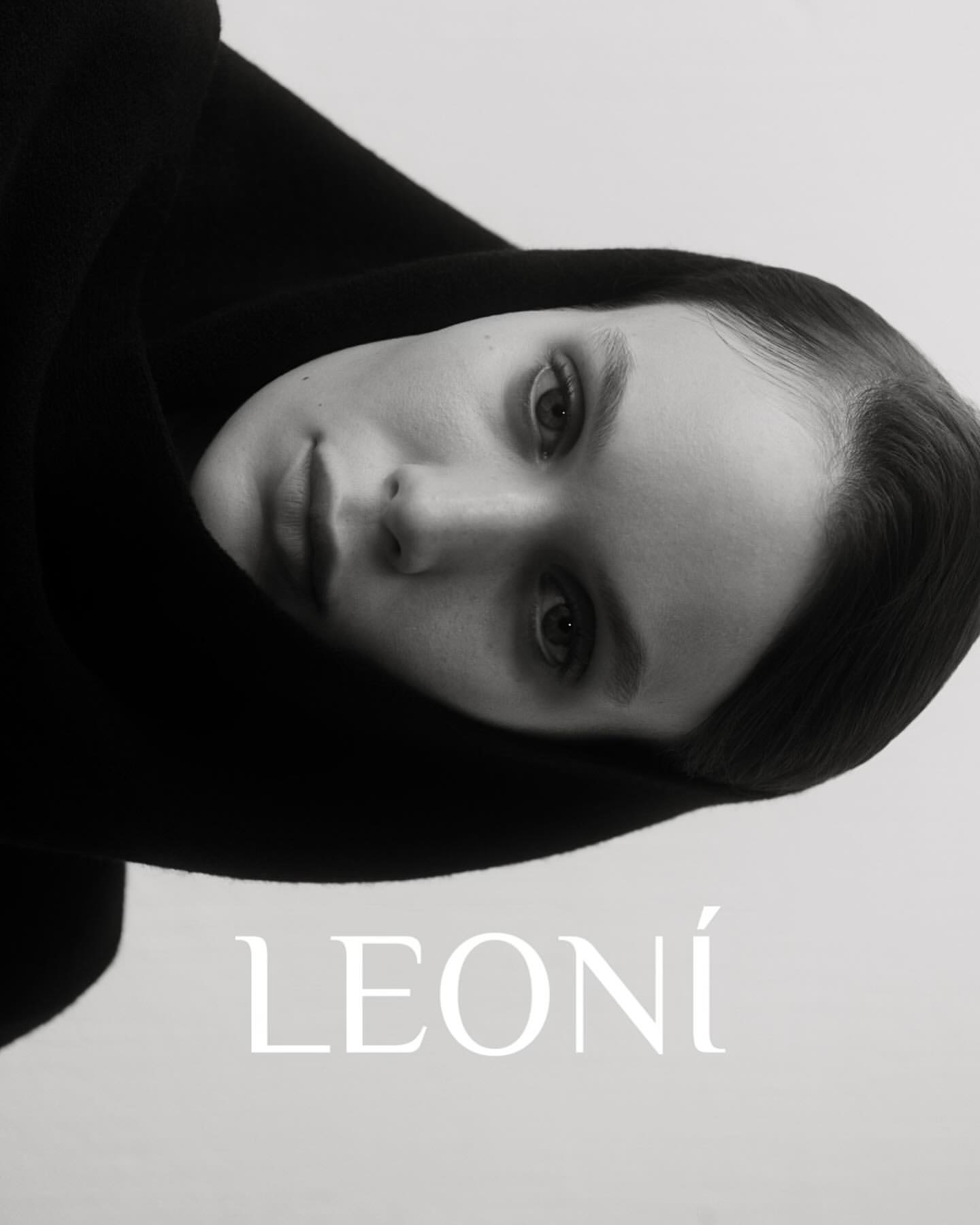 Monochromatic close-up of an adult female with fair skin and a neutral facial expression, wearing a black hooded garment and soft lighting.