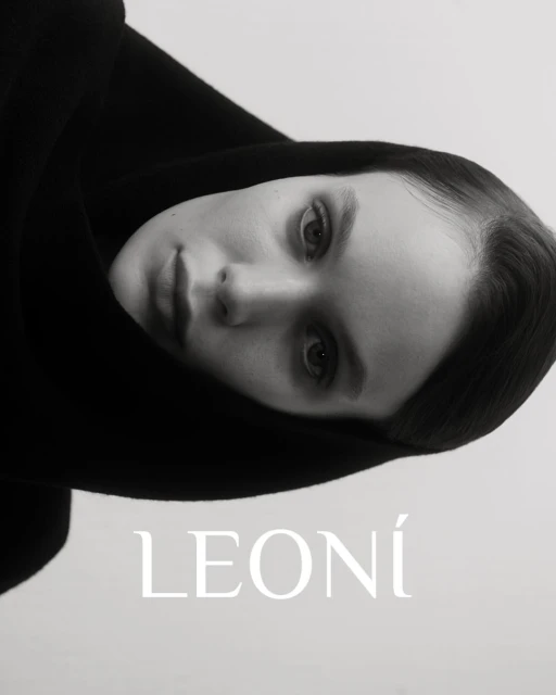 Monochromatic close-up of an adult female with fair skin and a neutral facial expression, wearing a black hooded garment and soft lighting.