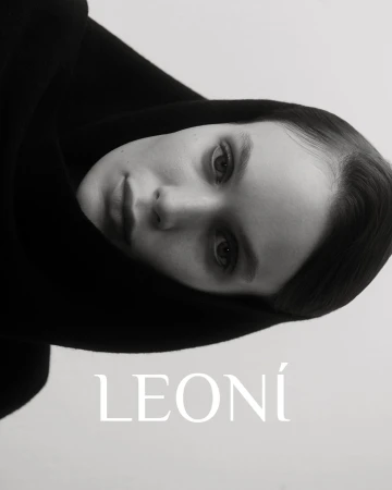 Monochromatic close-up of an adult female with fair skin and a neutral facial expression, wearing a black hooded garment and soft lighting.