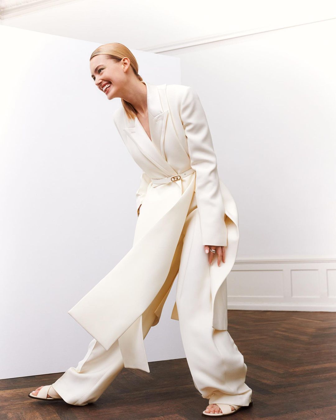Smiling woman in a cream-colored suit standing in an indoor setting. She has blonde hair and is wearing open-toed heeled shoes.
