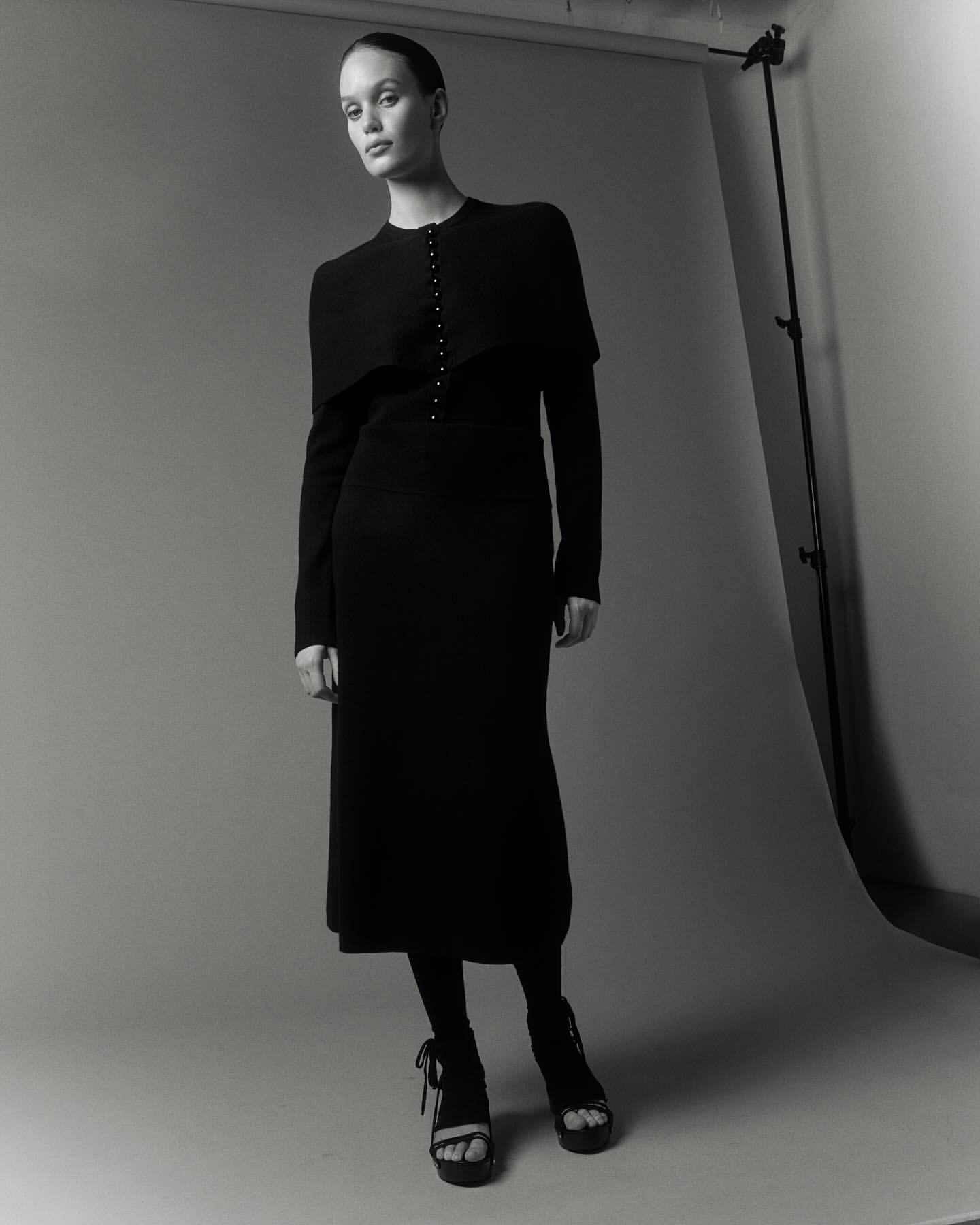 Monochrome studio shot of a woman in a black dress with matching tights and platform heels, posing against a neutral backdrop.