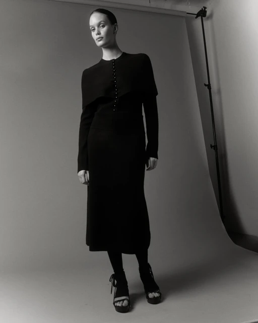 Monochrome studio shot of a woman in a black dress with matching tights and platform heels, posing against a neutral backdrop.