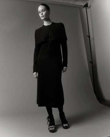 Monochrome studio shot of a woman in a black dress with matching tights and platform heels, posing against a neutral backdrop.