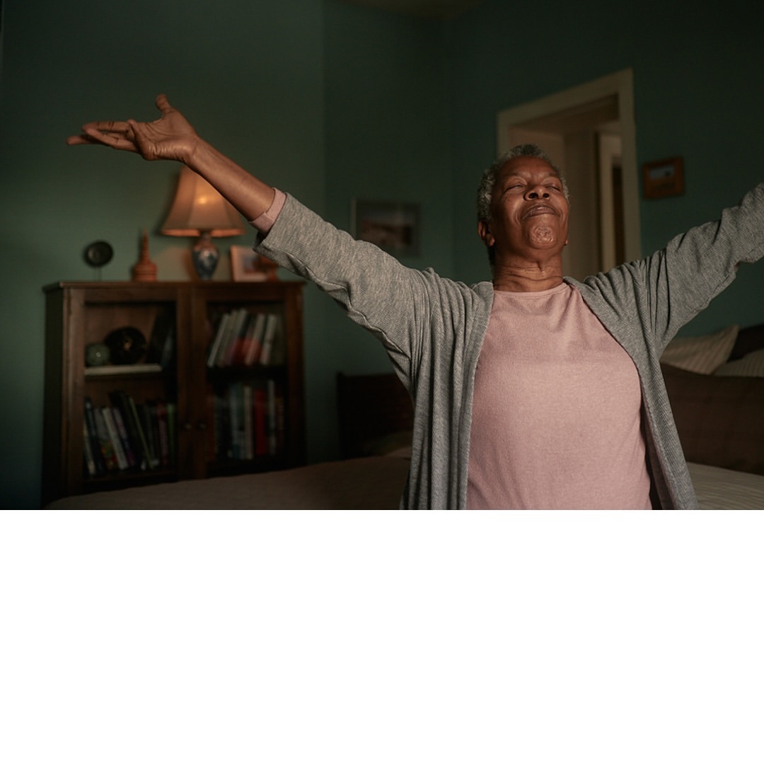 Older African-American woman with arms outstretched in bedroom. Gray cardigan, pink shirt, and soft, natural light.