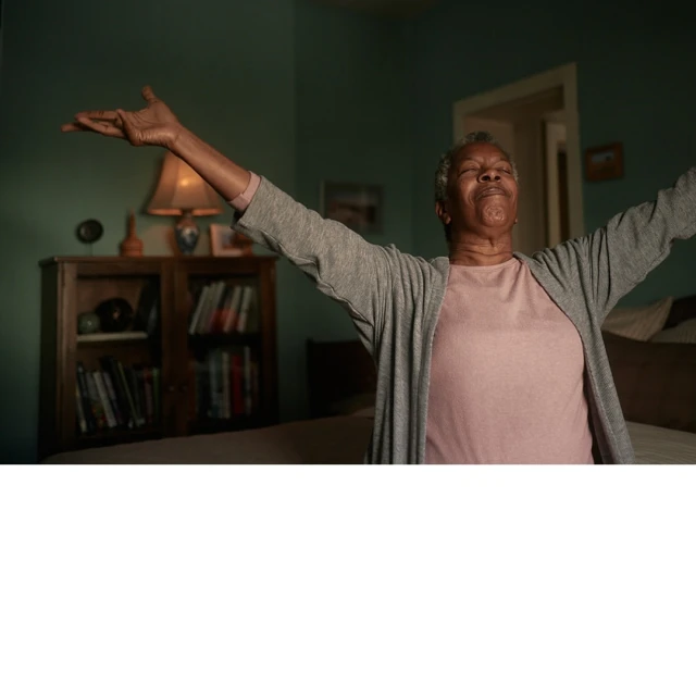 Older African-American woman with arms outstretched in bedroom. Gray cardigan, pink shirt, and soft, natural light.