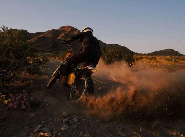 A motorcyclist wearing protective gear drives a dirt bike along a dirt path, kicking up dust as mountains fill the background.