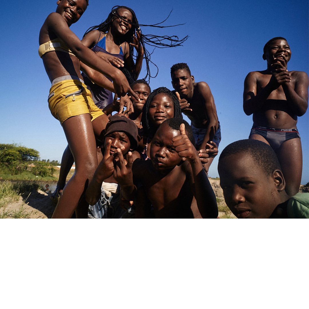 Group of African youth smiling and posing outdoors in swimwear, under a clear sky, captured in a candid, eye-level shot on a sandy beach.