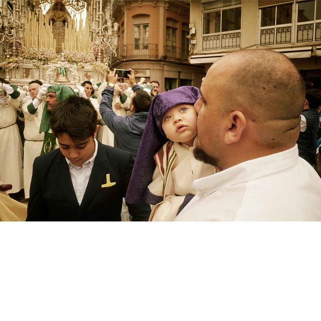 Father holding toddler in traditional religious attire, procession with hooded figures in background on urban street in daylight.