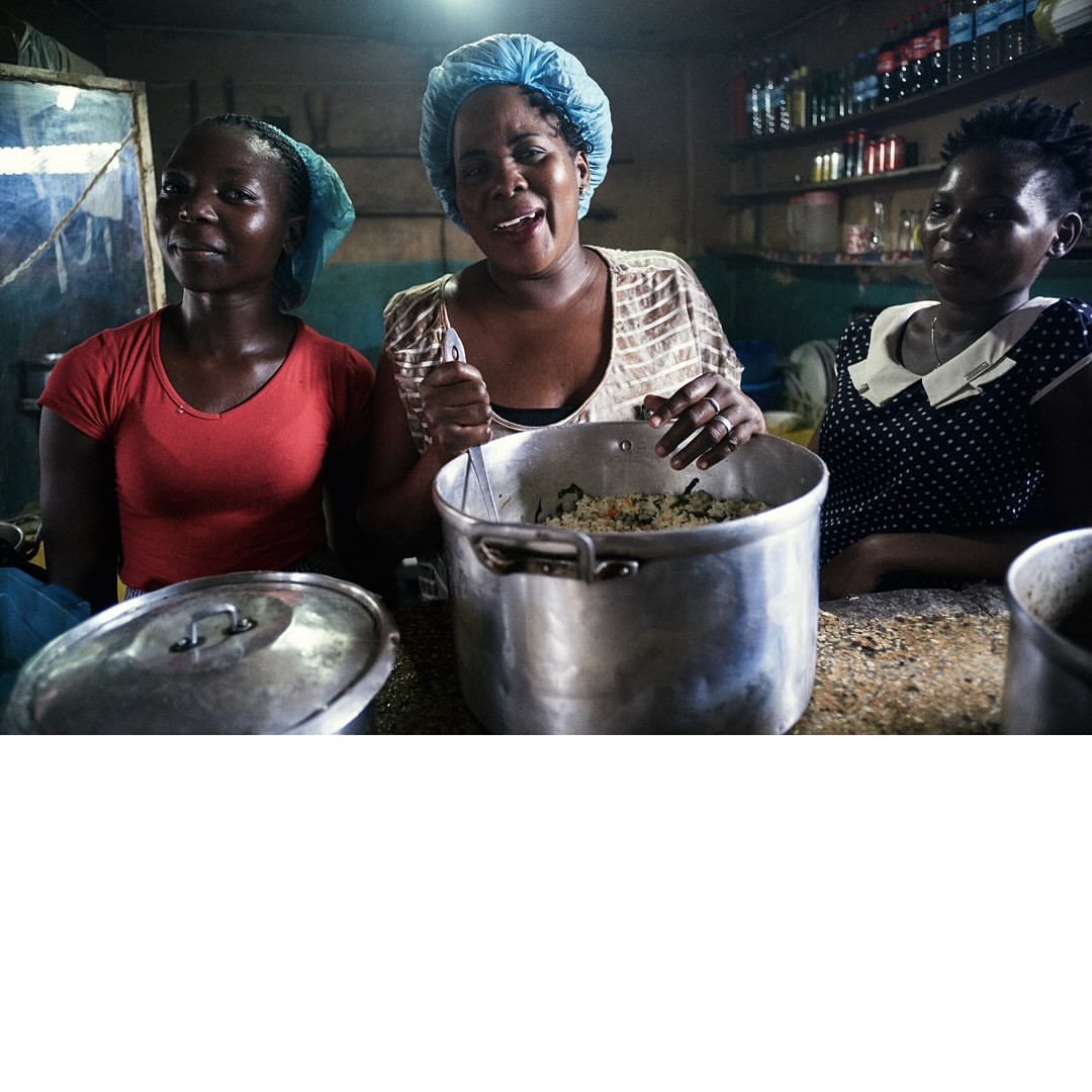 Three African women preparing food in a kitchen, wearing hairnets, with large pots, in an indoor setting, with natural light.