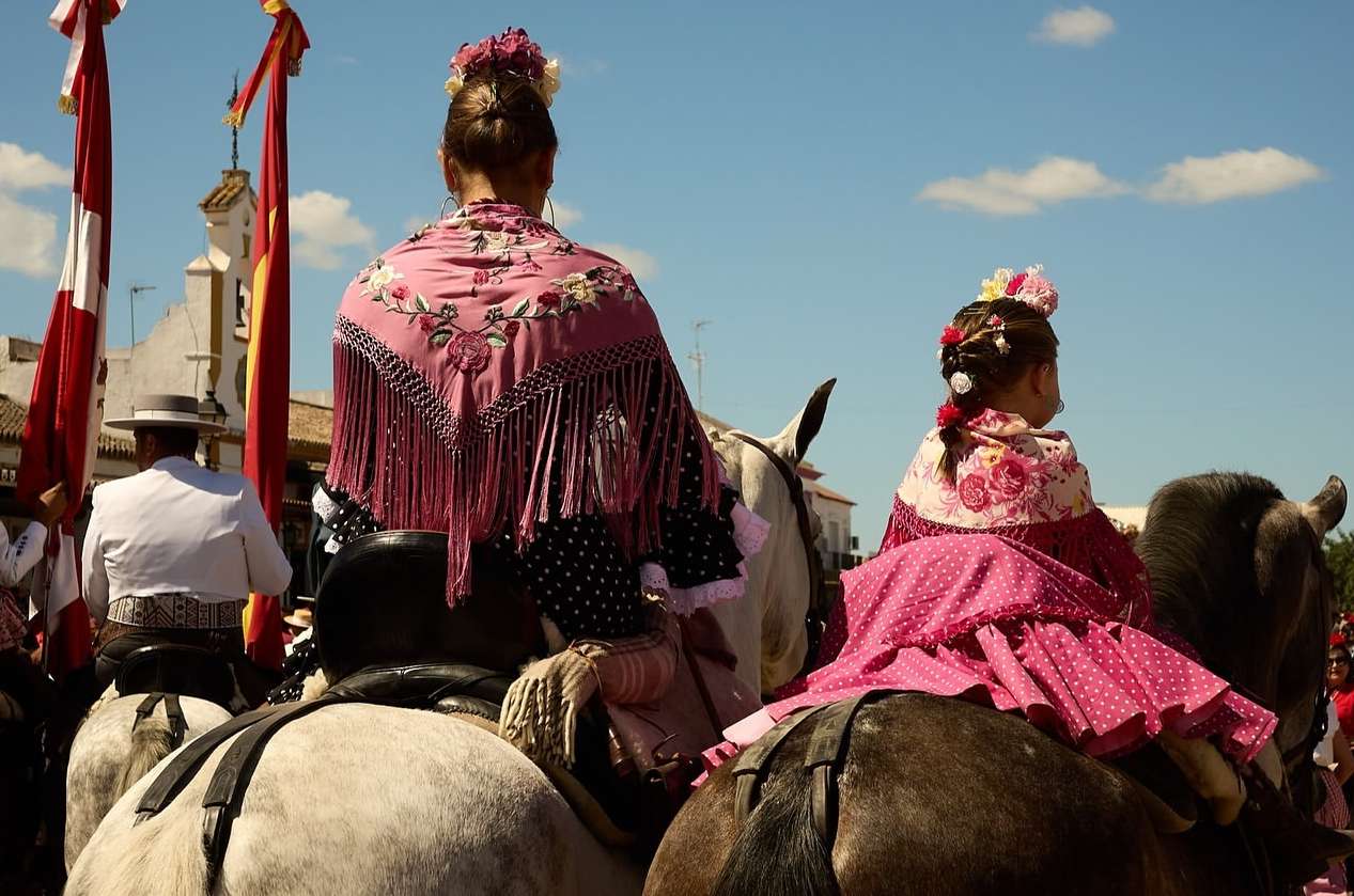 Two people in traditional dresses on horseback, viewed from behind, under a bright sky with buildings and flags in the background.