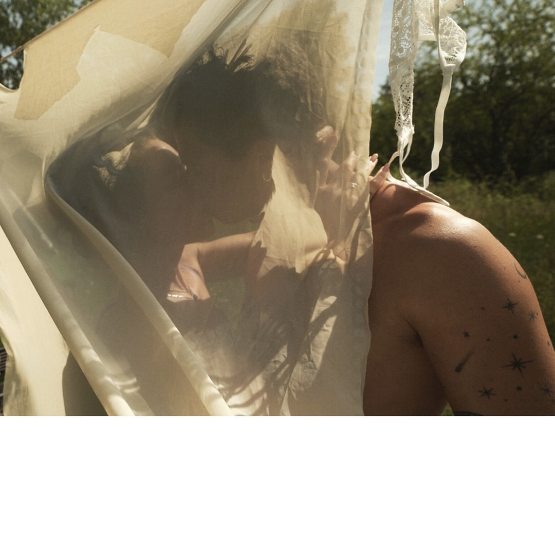 An artistic, intimate outdoor portrait of a topless adult with a constellation tattoo partially veiled by a sheer, textured fabric.