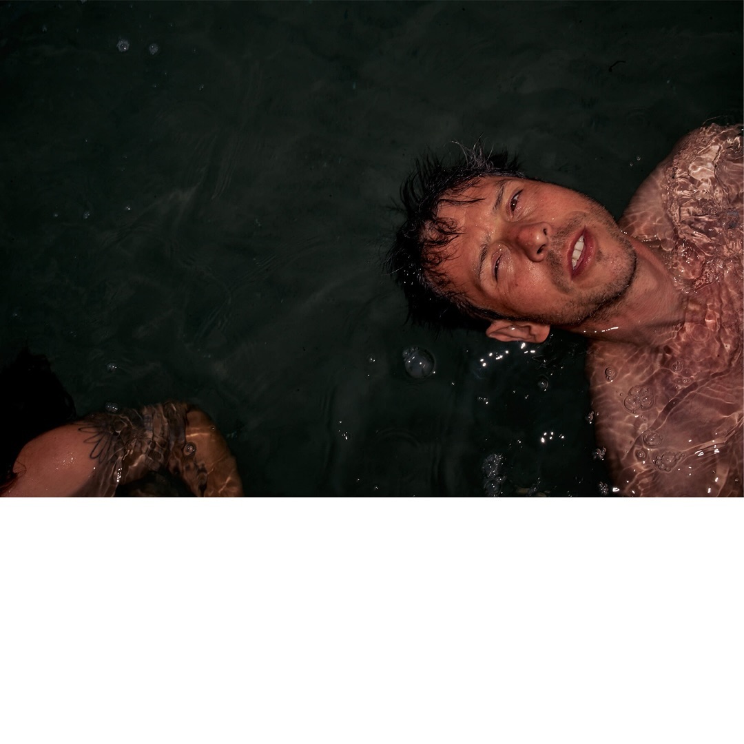 Man floats in dark water, partially submerged with an arm visible in the lower left; the scene is shadowy and atmospheric.