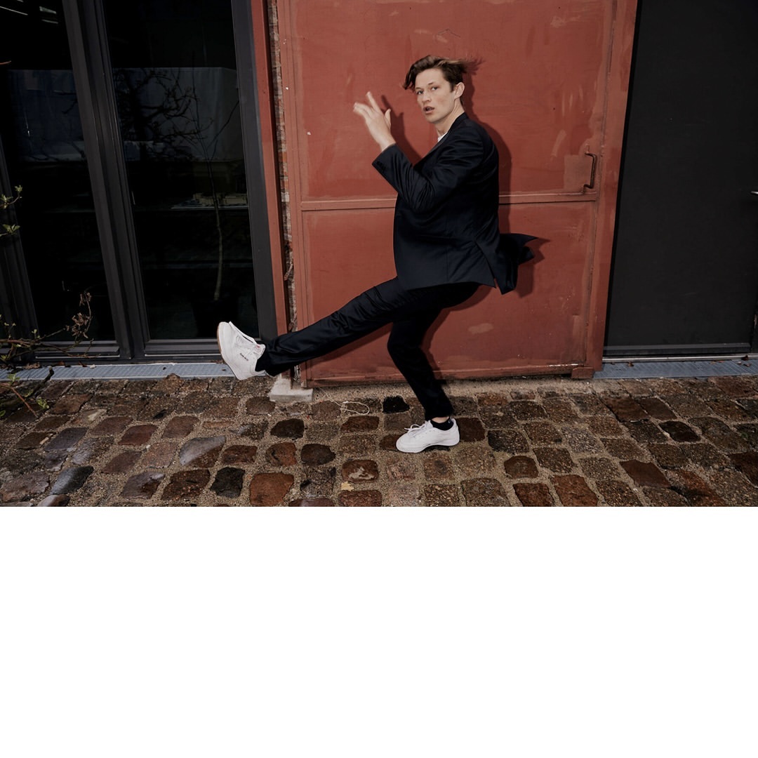 Young man in suit playfully posing with leg extended near red door and brick sidewalk, wearing white sneakers outdoors.