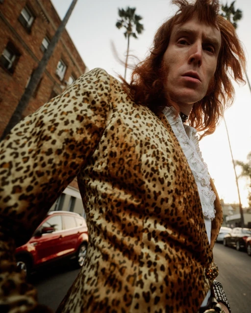 A man with red hair in an animal-print blazer stands on an urban street with cars and palm trees in the background.