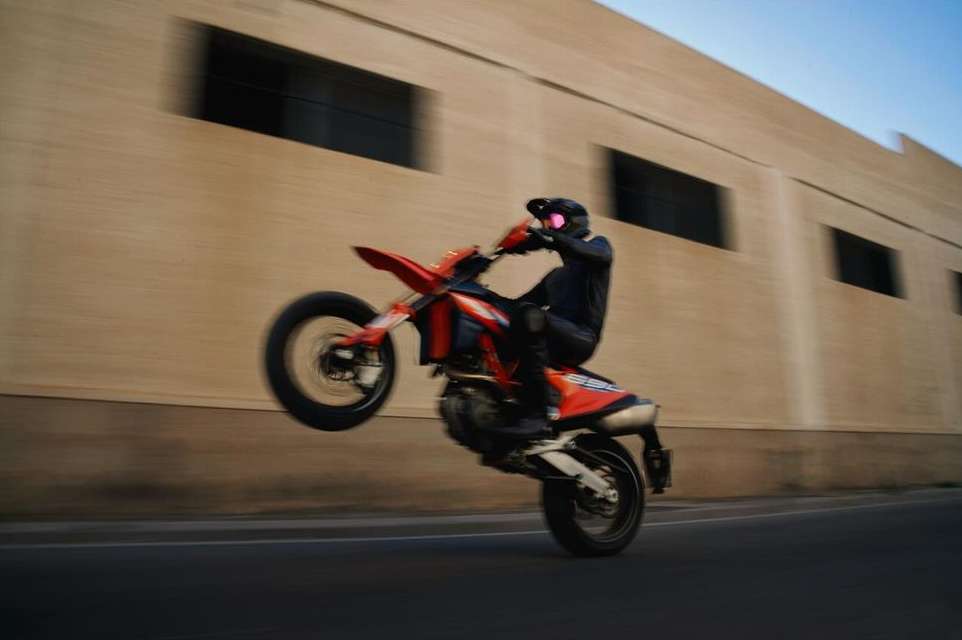 Motorcyclist performs a wheelie on an orange motorbike on a paved road with a beige building background on a sunny day.