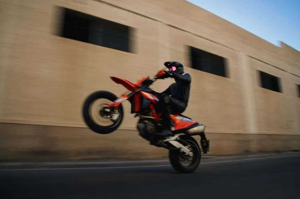 Motorcyclist performs a wheelie on an orange motorbike on a paved road with a beige building background on a sunny day.