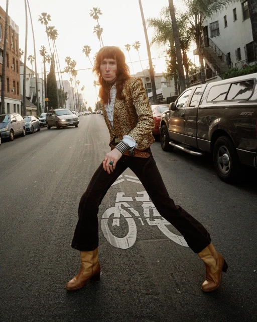 A long-haired person in patterned jacket and bell-bottoms strides across a street with palm trees and cars in the background at dusk.