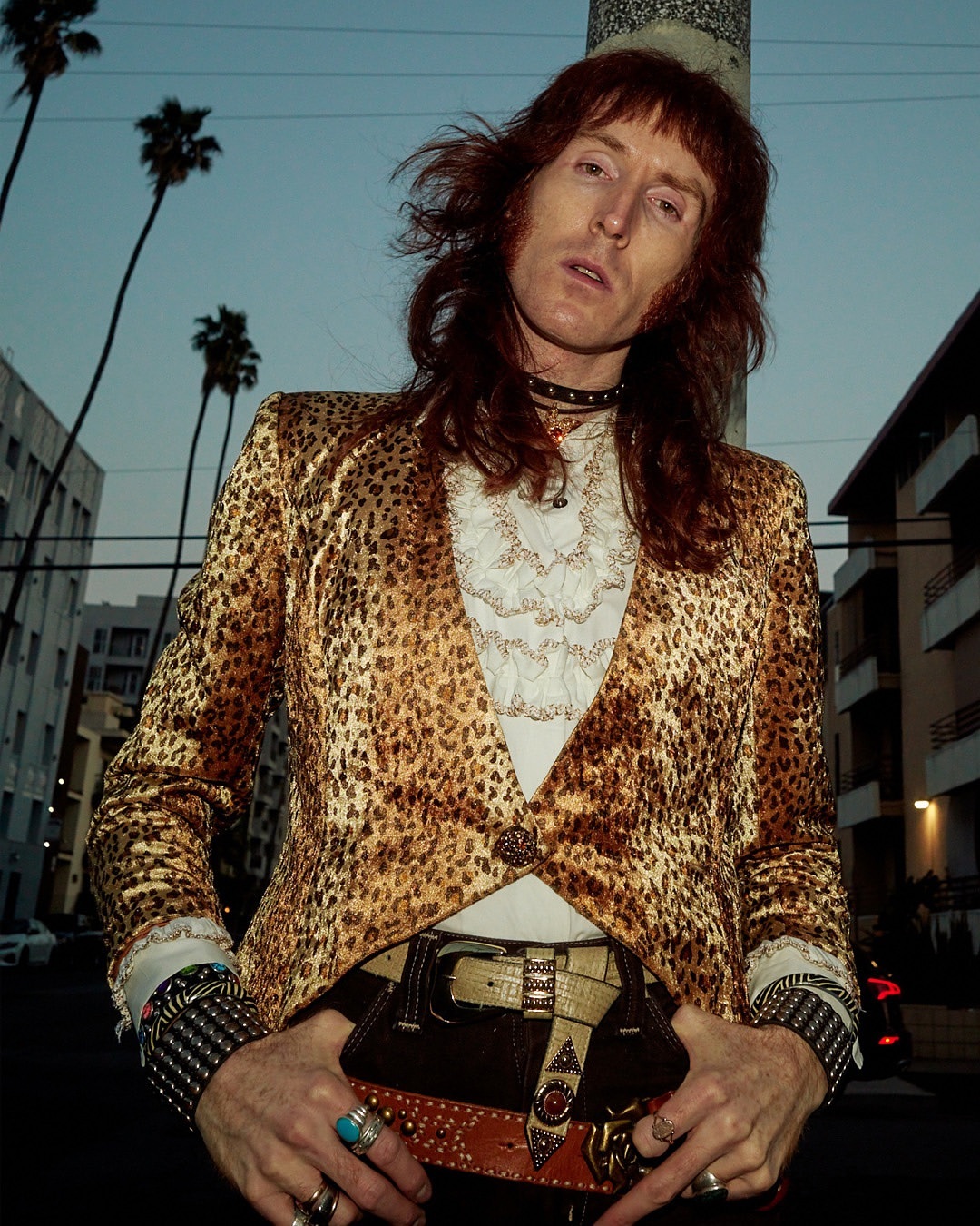 Adult male with long hair posing in a leopard print jacket and belt with palm trees and a building in the background on a city street.