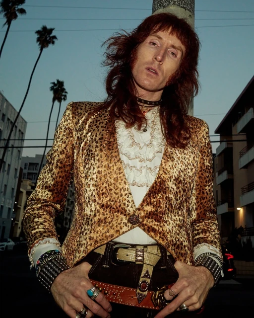 Adult male with long hair posing in a leopard print jacket and belt with palm trees and a building in the background on a city street.