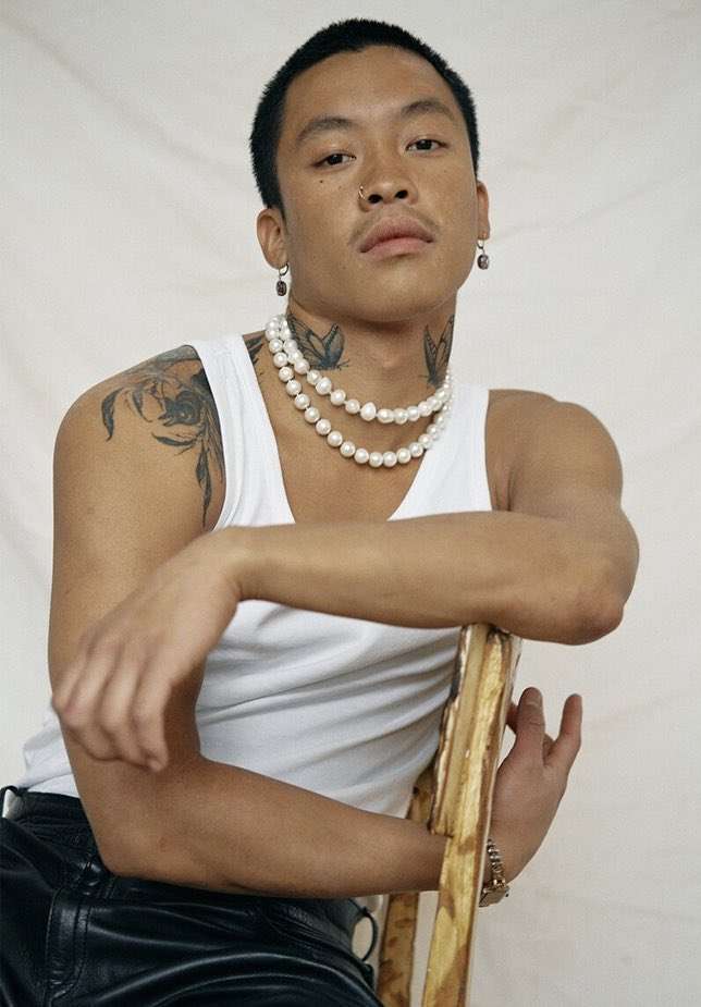 Asian man sits on gold chair in photo studio wearing tank top, black pants, pearl necklaces, and jewelry posing for camera.