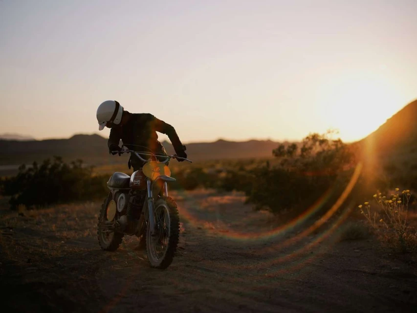 A motorcyclist pauses on a dirt road in a desert landscape at sunset, wearing a helmet and dark clothing next to a vintage motorcycle.