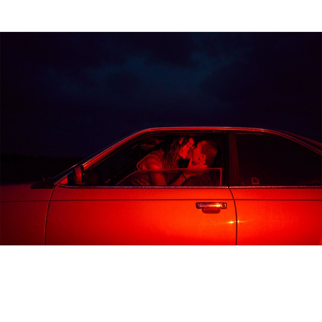 Side view of a kissing couple in a car at night, bathed in red lighting, suggesting a romantic or cinematic scene.