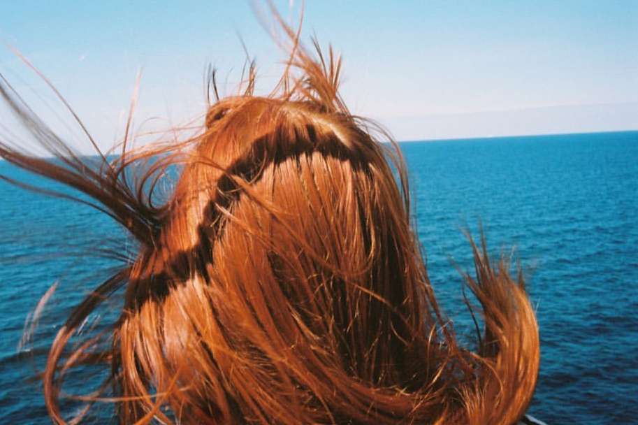 Back of a red-haired woman's head with the wind blowing her hair around her face, set against a backdrop of blue ocean and sky.