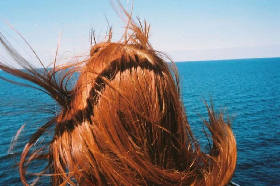 Back of a red-haired woman's head with the wind blowing her hair around her face, set against a backdrop of blue ocean and sky.