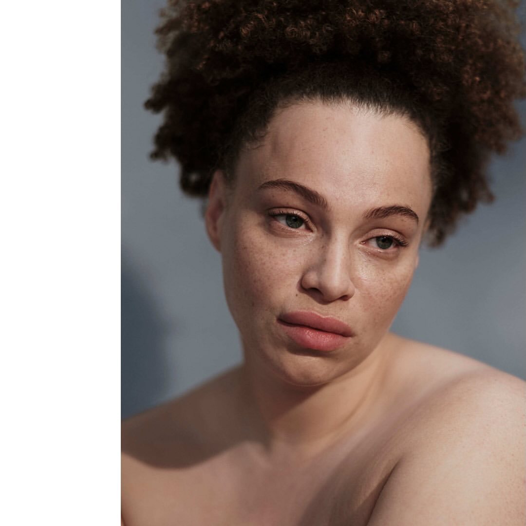 Close-up portrait of a light-skinned woman with curly dark hair against a muted grey background, showcasing beauty and natural aesthetic.