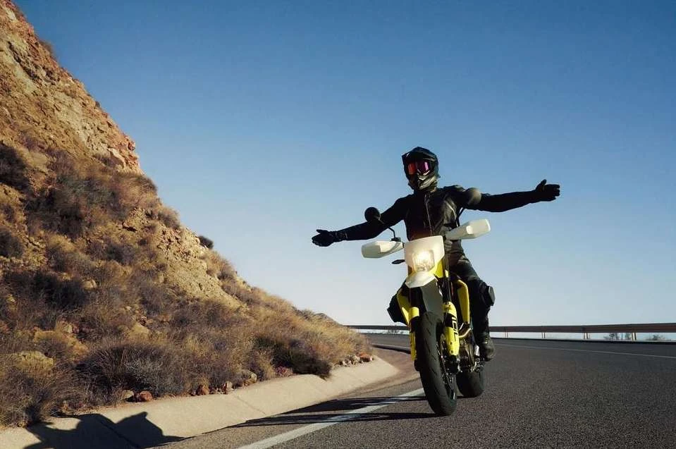 A motorcyclist wearing full protective gear rides a yellow motorcycle on an open road on a sunny day with a mountain backdrop.