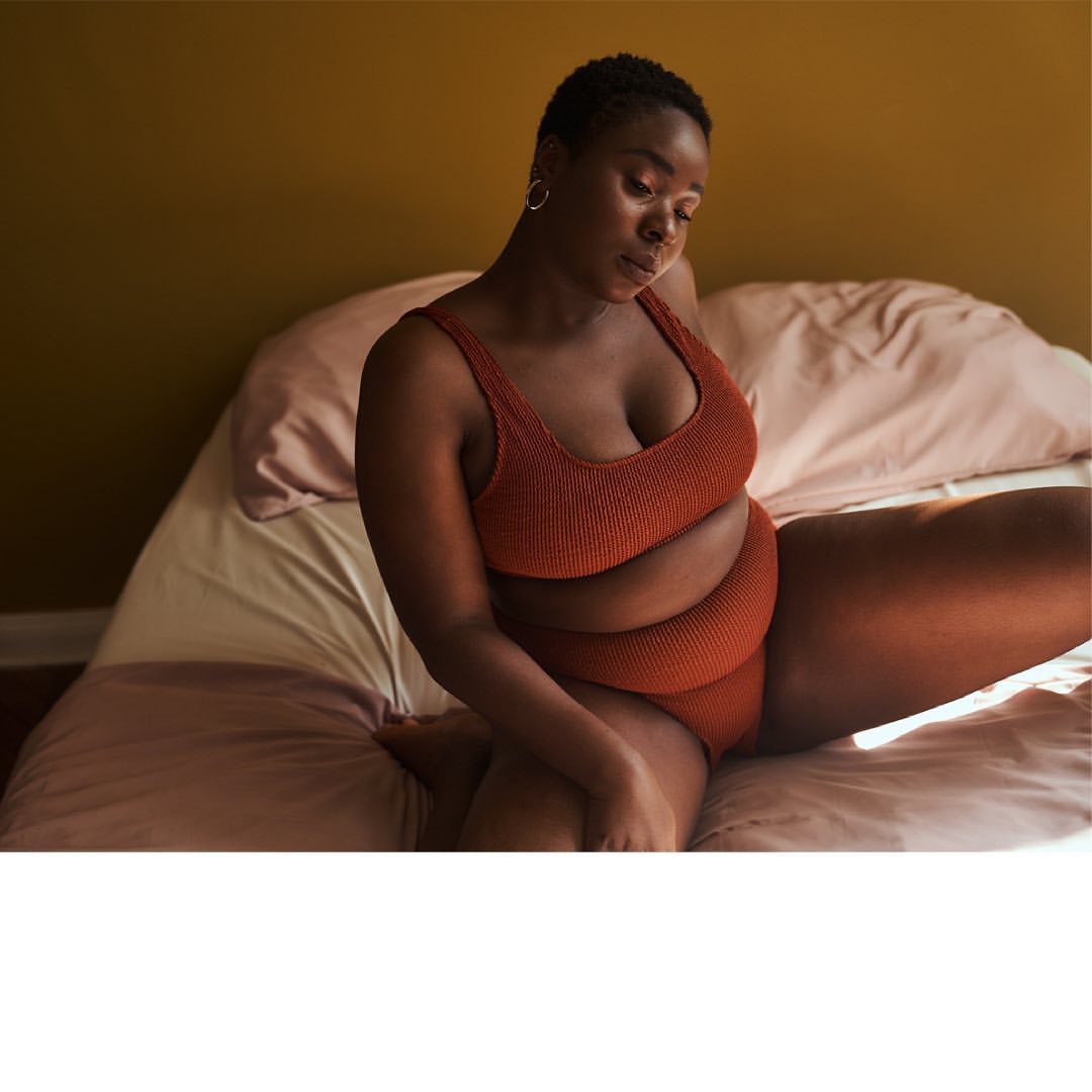 A plus-size African-American woman in a rust-colored ribbed two-piece sits on a bed with peach-colored sheets in a naturally lit room.