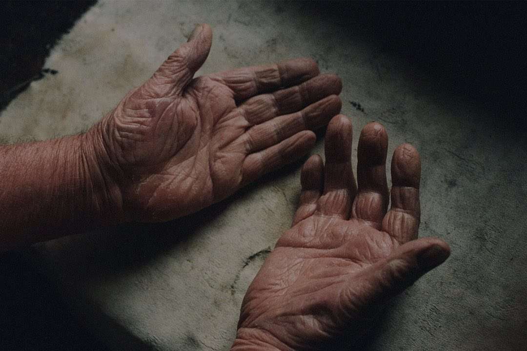Close-up photo of elderly male hands with wrinkles, conveying age, texture, and detail against a blurred, neutral background.